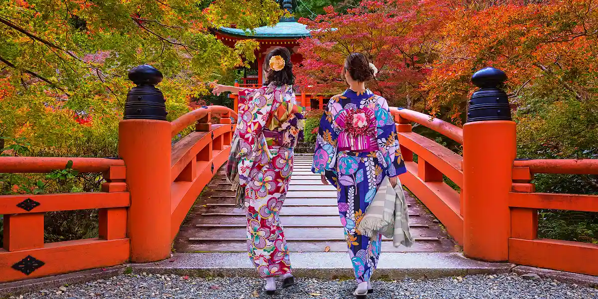 Two travellers in kimono crossing a red temple bridge in autumn